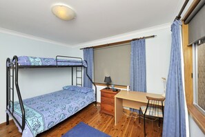 Ein Stockbett mit einem gemusterten Bettbezug, ein Holztisch mit Stuhl, eine Kommode und ein Fenster mit Vorhängen.