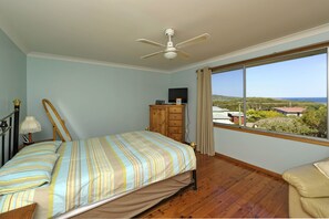Een slaapkamer met een bed, een houten dressoir, een plafondventilator en een raam met uitzicht op zee.