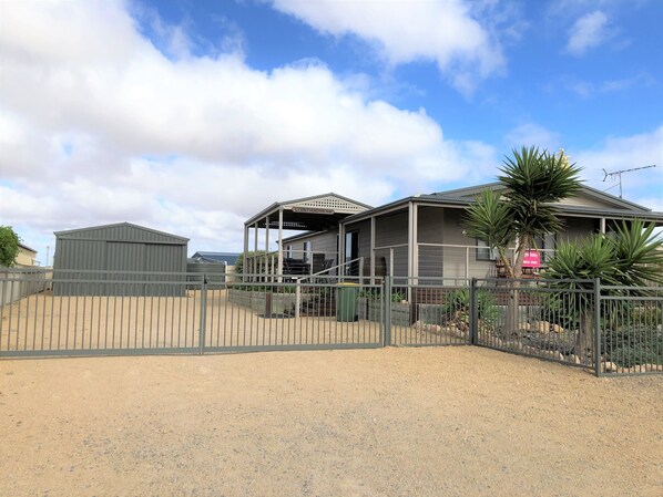 A property with a fenced driveway, a covered outdoor area, and a building with a sign indicating a sale.