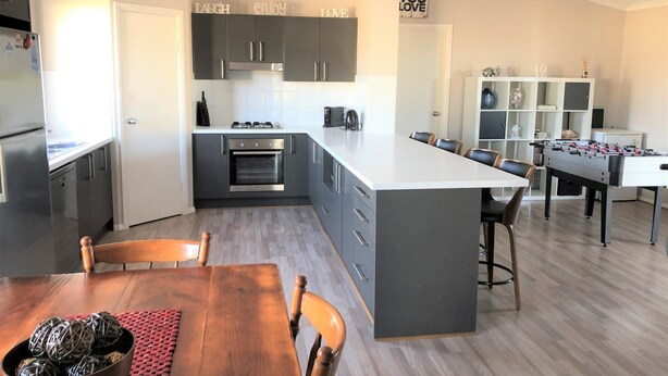 A modern kitchen with grey cabinets, a central island, and a dining area with a wooden table and chairs. - Livin' The Dream ~ Spacious  ~  Off Peak Special! WIFI Available (Point Turton)