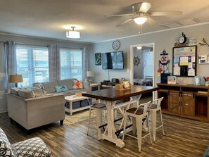 Un salon confortable avec un canapé, une table basse et une table à manger avec des chaises. Un téléviseur est fixé au mur, il y a un ventilateur de plafond et un tableau d’affichage où sont accrochés divers objets.