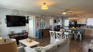 A living room with a sofa, a coffee table, a TV, and a dining area with chairs and a kitchen in the background. - Experience beachfront living at Sandy Bottoms in Holden Beach, NC—perfect for families! (Holden Beach)