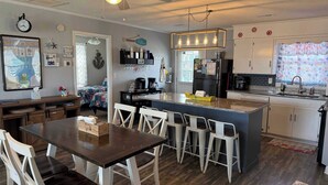 A modern kitchen with a central island, white bar stools, and a dining table. - Experience beachfront living at Sandy Bottoms in Holden Beach, NC—perfect for families! (Holden Beach)