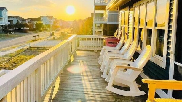 A wooden deck with white rocking chairs, a view of the sunset, and a building with large windows. - Experience beachfront living at Sandy Bottoms in Holden Beach, NC—perfect for families! (Holden Beach)