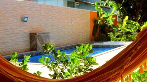 Una hamaca sobre una piscina, con un muro de ladrillo y follaje al fondo.