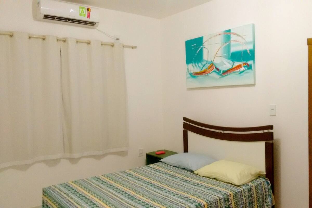 A bedroom with a bed, a nightstand, a wooden wardrobe, a painting on the wall, and an air conditioning unit.