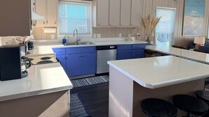 A modern kitchen with white cabinets, blue accents, a dishwasher, and a large island with bar stools. - Enjoy oceanfront views at How Sweet The Sound in Holden Beach, NC; perfect for families. (Holden Beach)
