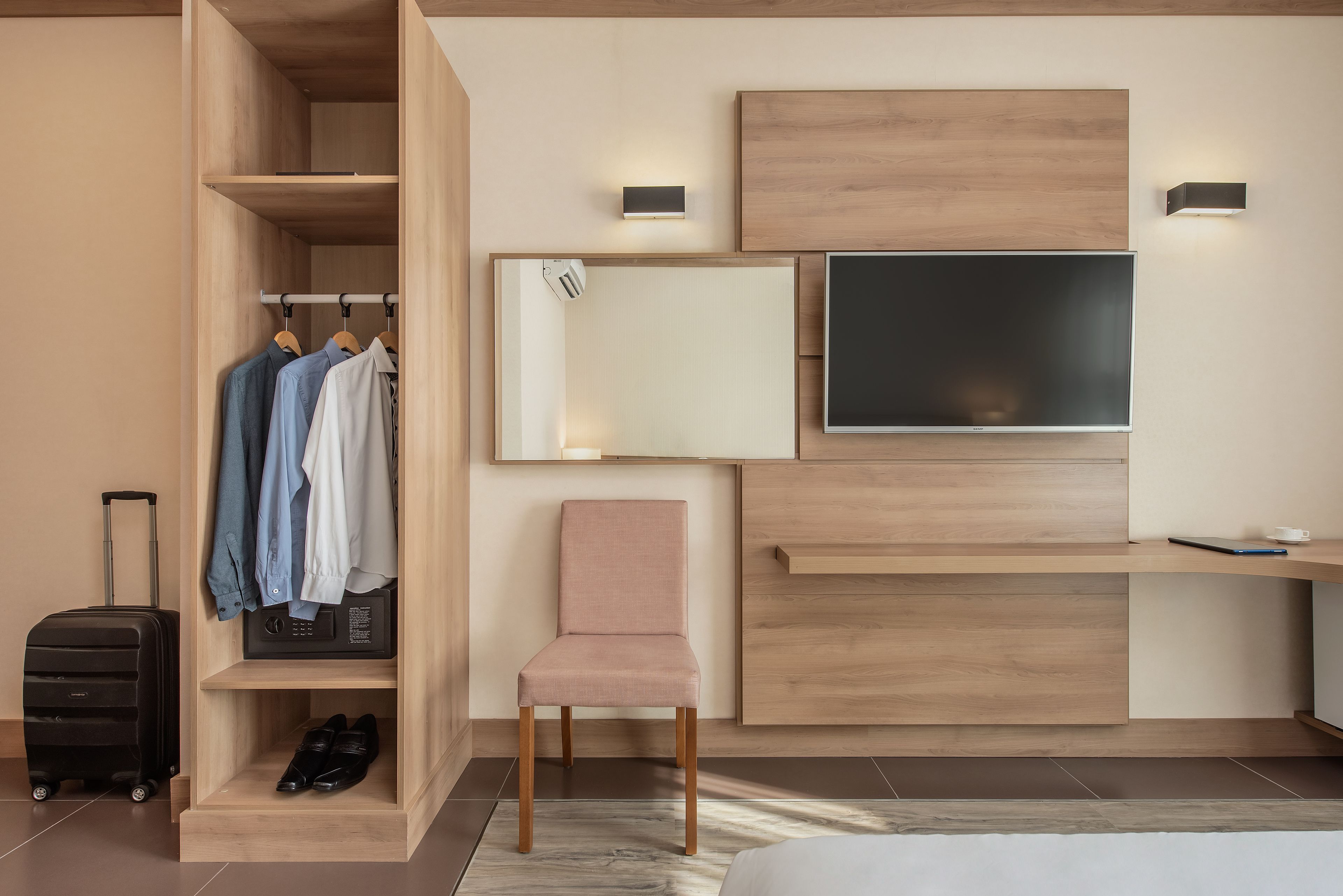 A modern hotel room with a wooden wardrobe, a flat-screen TV, a pink chair, and a suitcase.