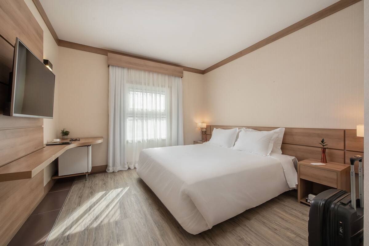 Quarto de hotel com uma cama grande, televisão na parede, escrivaninha com cadeira, mesa de cabeceira com abajur e mala no chão.