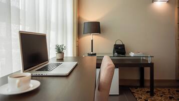 Habitación de hotel moderna con una laptop sobre un escritorio de madera, una pequeña planta en maceta, una lámpara y un bolso de mano sobre una mesa de vidrio.