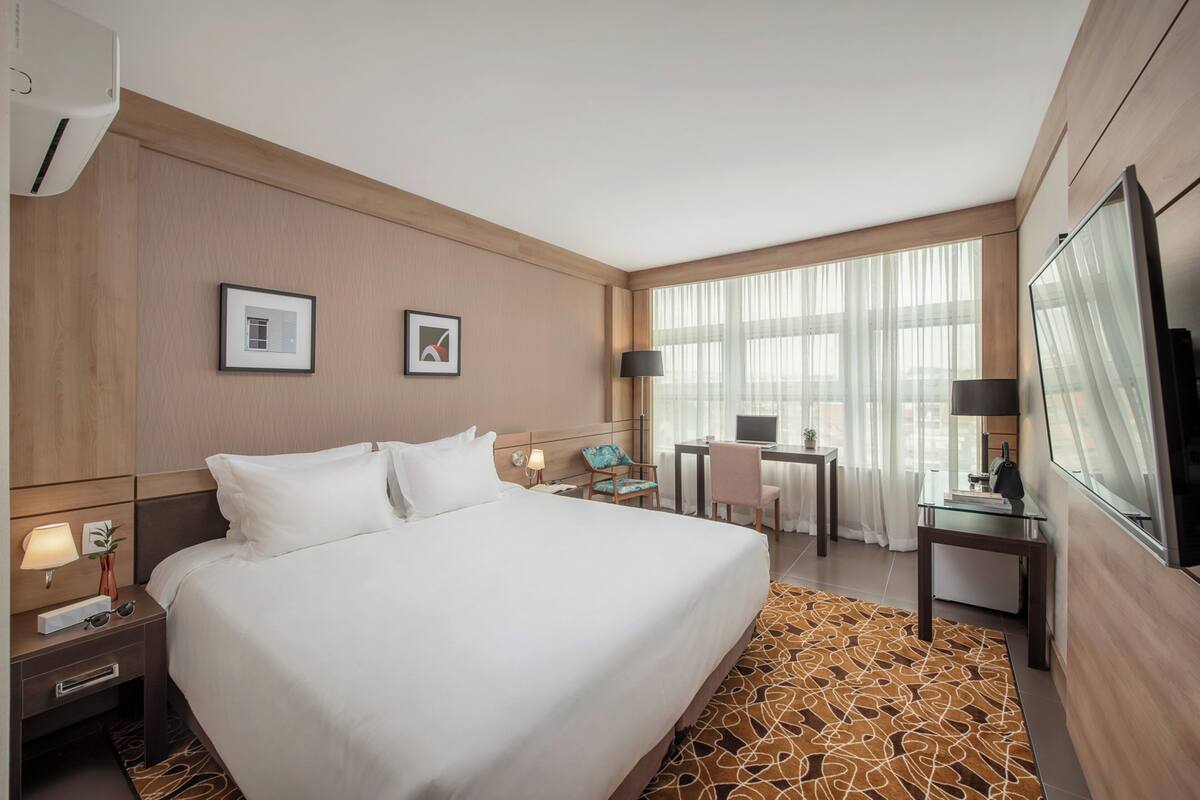 Quarto de hotel com uma cama grande, uma escrivaninha com um computador, uma cadeira e uma televisão.