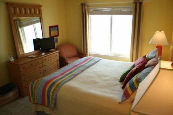 A hotel room with a bed, a television on a dresser, a chair, and a window with curtains. - Experience oceanfront bliss at Next 2 Paradise, a cozy 2BR condo in Holden Beach. (Holden Beach)