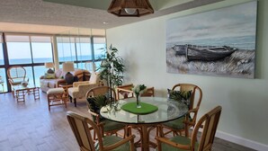 Un'area pranzo con un tavolo rotondo in vetro, circondato da sedie in vimini con cuscini verdi, un lampadario in vimini a sospensione e un dipinto di una barca appeso al muro.