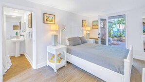Une chambre à coucher comprenant un lit, des tables de chevet, une commode, une fenêtre donnant sur un espace verdoyant, et une salle de bain avec un lavabo et un miroir.