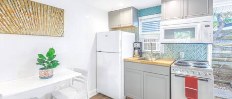 Una cocina compacta con armarios blancos, un refrigerador blanco, una estufa y un microondas. Hay una mesa pequeña con sillas y una planta en maceta.