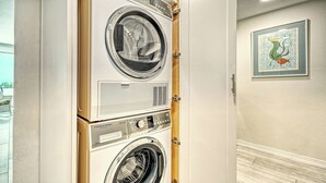 Un appareil combiné lave-linge et sèche-linge, avec un panneau de commande, une œuvre d’art encadrée accrochée au mur et une vue sur l’extérieur.