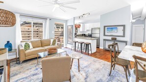 A spacious living area with a sofa, coffee table, and a dining table with chairs. There is a kitchen area with a bar and stools. - Gertrude's Siesta | Top Location! 1-min to Best Beaches, Private Patios, Gate to Village! (Siesta Key)