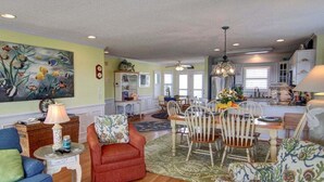 A spacious living room with a dining area, a kitchen, and a large painting on the wall. - Turtle Cove is a spacious 4-bed home in Holden Beach, perfect for 9 guests. (Holden Beach)