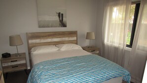 A neatly made bed with a striped bedspread, two bedside tables with lamps, a wooden headboard, and a window with sheer curtains. - The Catch  ~ short walk to beach! Dog Friendly  - Off Peak Special! (Stansbury)