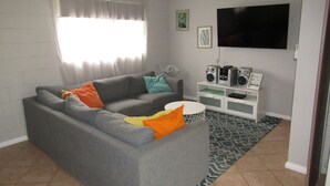A living room with a grey sofa, colorful cushions, a white entertainment unit with speakers, a flat-screen TV, and a patterned rug. - The Catch  ~ short walk to beach! Dog Friendly  - Off Peak Special! (Stansbury)