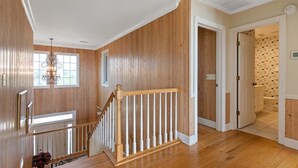 A wooden interior with a staircase, a chandelier, and a view of a bathroom with patterned wallpaper. - Cypress Hall: Stunning 6-Bedroom, Oceanfront Retreat in Holden Beach! (Holden Beach)
