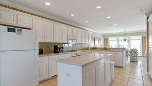 A kitchen with white cabinets, a refrigerator, a coffee maker, and a dining area with chairs. - Cypress Hall: Stunning 6-Bedroom, Oceanfront Retreat in Holden Beach! (Holden Beach)