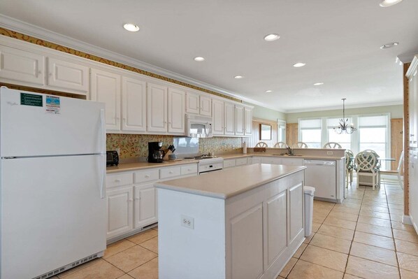 A kitchen with white cabinets, a refrigerator, a coffee maker, and a dining area with chairs. - Cypress Hall: Stunning 6-Bedroom, Oceanfront Retreat in Holden Beach! (Holden Beach)