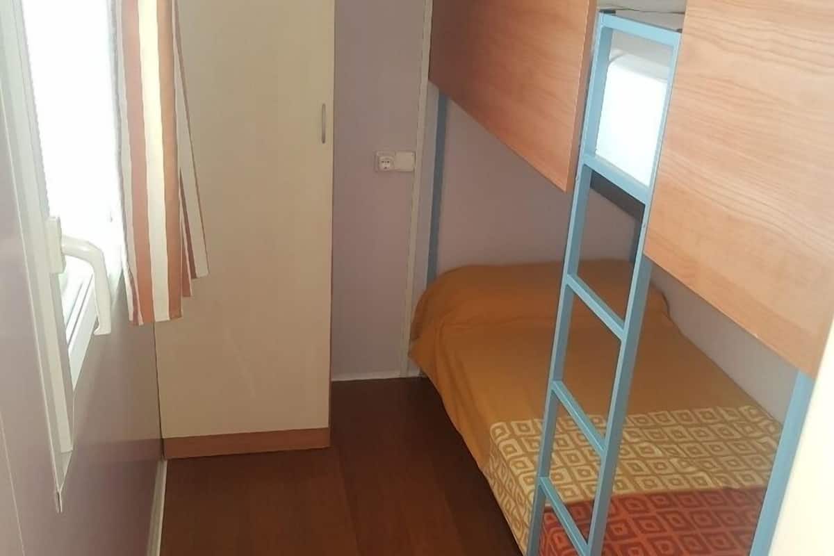 Une petite pièce étroite, avec un lit superposé, une porte et une fenêtre avec des rideaux.