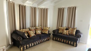 Un salón con dos sofás oscuros, cojines decorativos estampados y cortinas color beige.