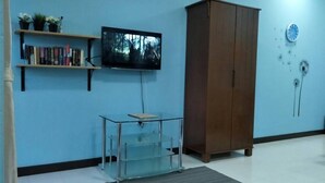 A room with a TV mounted on the wall, a bookshelf with books and plants, a wooden cabinet, and a glass table with a rug. - Triple B's Staycation - Simple and Comfy (Petaling Jaya)