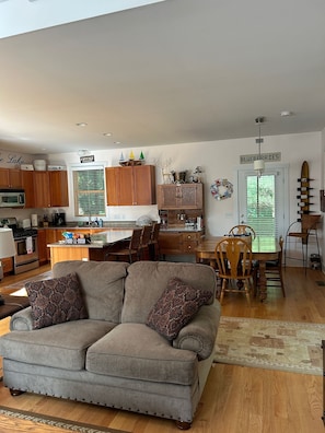 A cozy living room with a grey sectional sofa, wooden dining table, and chairs. A kitchen with wooden cabinets and a window is visible in the background.