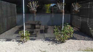 Un patio couvert avec une table en bois et des bancs, entouré d’une clôture en métal et d’un sol en gravier.