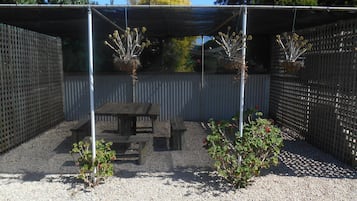 A covered patio area with a wooden table and benches, surrounded by a metal fence and gravel ground.