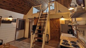 A rustic kitchen with wooden beams, a stove, and a ladder leading to a loft area. - Black Hills Farmhouse, Cabin #3 (Custer)