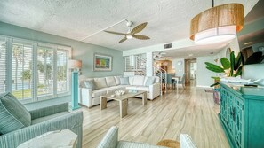 A spacious living room with a white sofa, a coffee table, and a dining area with a table and chairs. There is a staircase leading to another level, and a large window with blinds.