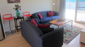 Ein Wohnzimmer mit einem blauen Sofa, roten und schwarzen Kissen, einem kleinen Tisch mit einer Schale, einer Vase mit Blumen und einem roten Sessel.