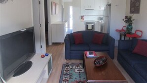 Ein Wohnzimmer mit Fernseher, Sofa und Couchtisch. Im Hintergrund ist eine Küche zu sehen.