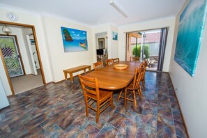 A dining area with a large wooden table, chairs, and a bench. There is a painting on the wall and a view of the outdoors through a glass door. - Government Rd 63 - Shoal Bay (Shoal Bay)
