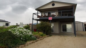 A two-story building with a balcony, a small garden with white flowers, and a clear sky.