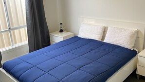 A neatly made bed with a blue bedspread and white pillows in a room with a window and a nightstand with a lamp.