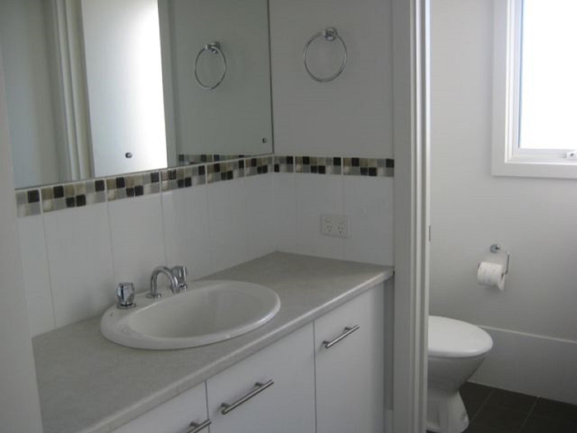 A bathroom with a white sink, mirror, and toilet. There is a towel ring above the mirror and a small window above the toilet.