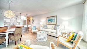 A modern living room with a dining area, a kitchen in the background, and a large window. - LaPlaya 101A - Step out to the beach.  Light and bright end unit. (Longboat Key)
