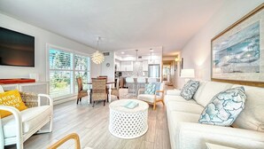 A modern living room with a sofa, chairs, a coffee table, and a dining area with a kitchen in the background. - LaPlaya 101A - Step out to the beach.  Light and bright end unit. (Longboat Key)
