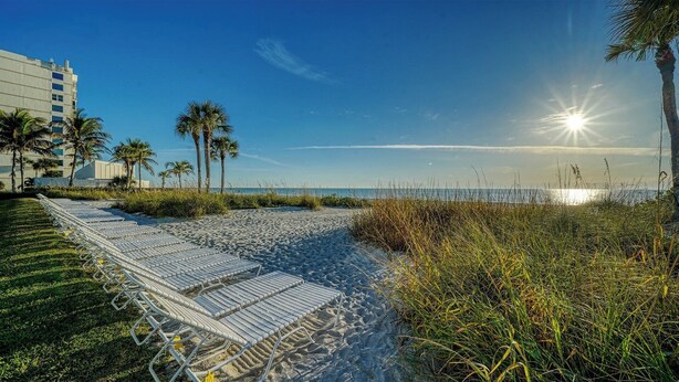 A beach with sunlit sand, palm trees, and a hotel in the background. - LaPlaya 102E-Tropical garden view near the pool! (Longboat Key)