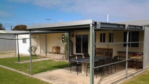 A covered outdoor patio with a table and chairs, a small garden area, and a building in the background.