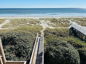 Une vue sur la plage, avec une promenade en bois qui mène au rivage, le tout entouré d’une végétation luxuriante.