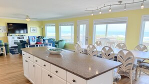 A kitchen with a granite countertop, white cabinets, and a dining area with white chairs and a table. - Experience oceanfront bliss at Summer Sandcastle, a spacious 5BR home in Holden Beach! (Holden Beach)