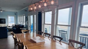 Ruime eetruimte met een houten tafel en stoelen, grote ramen met uitzicht op het strand, en een plafondventilator.
