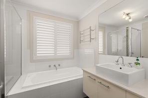Een badkamer met een bad, een wastafel, een spiegel en een raam met jaloezieën.