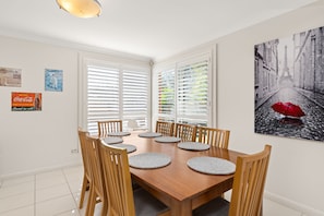 Een eetkamer met een houten tafel en stoelen, witte wanden en een schilderij van een regenachtige straat.
